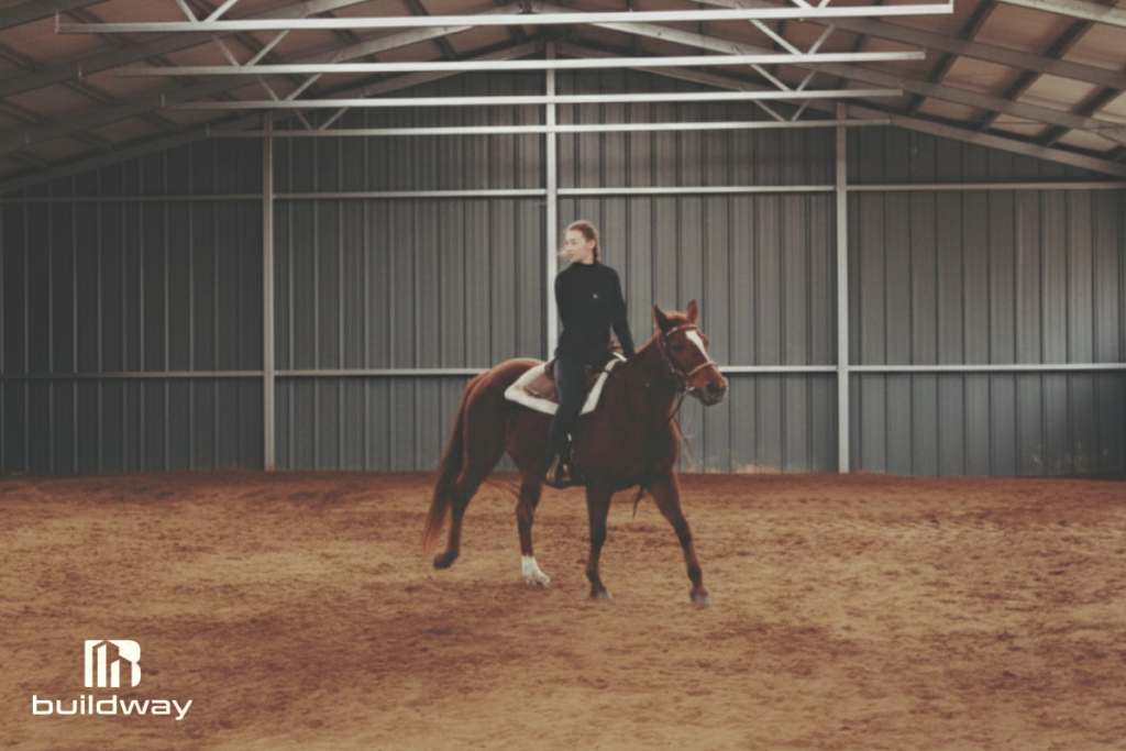 Equestrian rider practicing on a horse inside a spacious indoor agricultural riding arena with steel framing and dirt flooring, designed by Buildway.