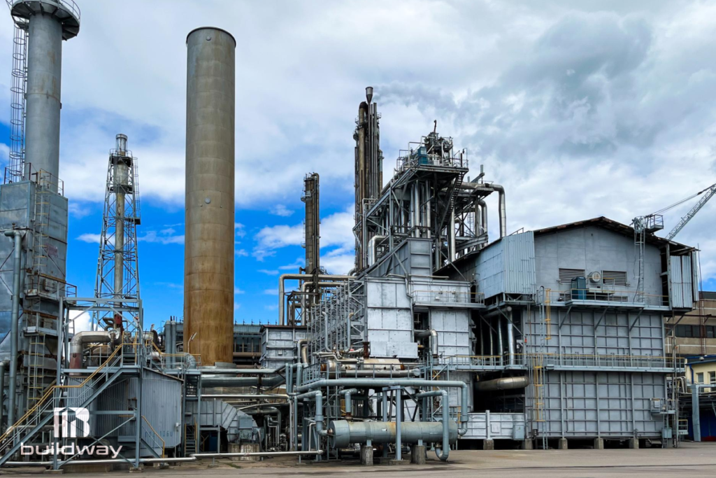 Complex industrial manufacturing plant with tall chimneys, metal pipelines, and structural frameworks under a partly cloudy sky, built by Buildway.