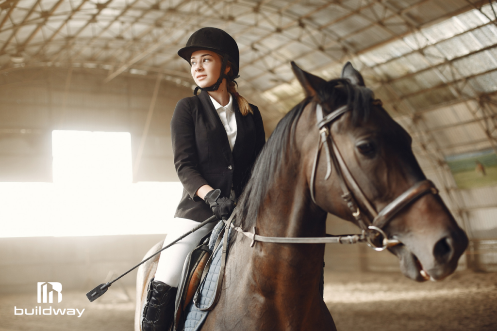 Equestrian rider on a horse inside a bright indoor agricultural riding arena with a high arched steel roof, designed by Buildway.
