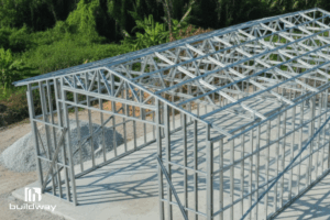 Steel I-beam building framework by Buildway showing a light-gauge structural skeleton with roof trusses and wall framing under construction on a concrete foundation, surrounded by greenery.