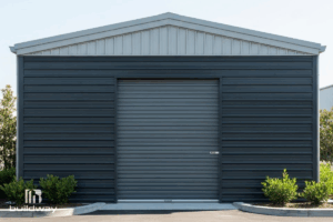 Modern I-beam steel building by Buildway featuring dark gray exterior wall panels, a central roll-up door, and a gable roof, with small green shrubs along the front.