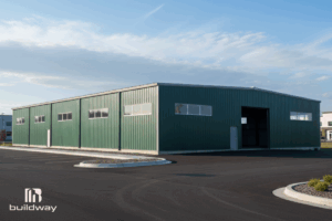 Large green I-beam steel structure by Buildway with multiple windows and a wide entrance, situated on a paved lot under a clear sky.