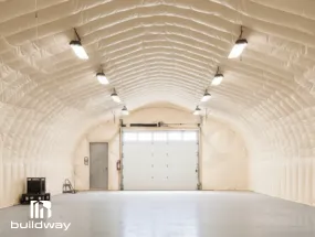 Interior of an insulated Quonset building by Buildway featuring white spray-foam insulation, overhead lighting, and a large garage door at the far end.