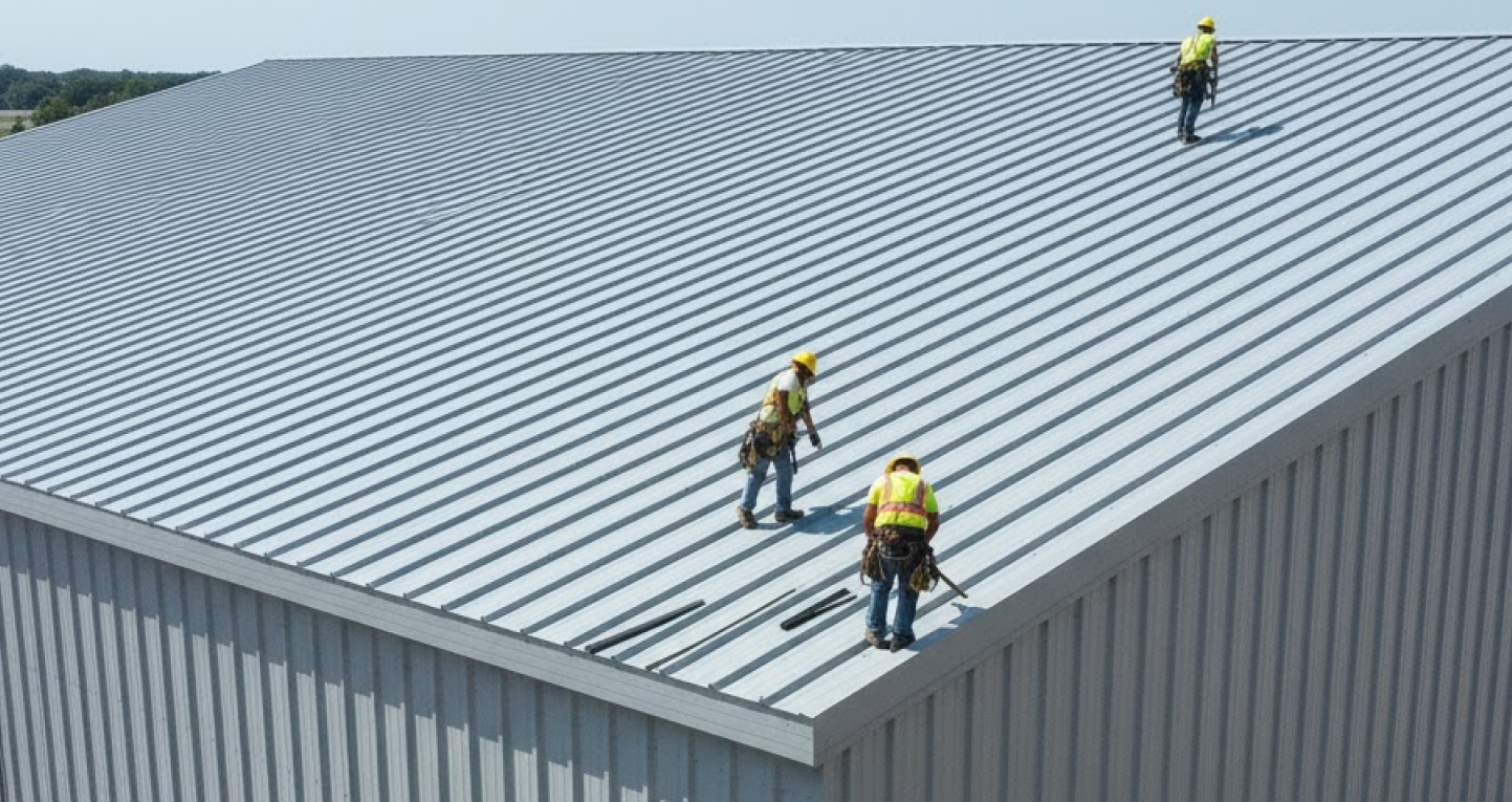 Construction workers installing metal roof panels on a steel building, with completed wall panels and trim package visible along the edges for sealing and aesthetics.