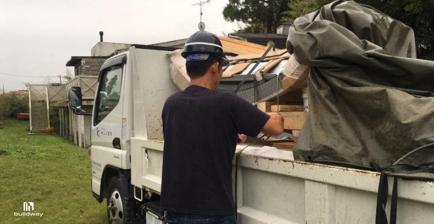 Why Your Steel Building Cost Is Higher Than Expected? (Full Breakdown With Hidden Fees) 6 Construction worker in a hard hat inspecting a delivery truck loaded with building materials, including wooden pallets and covered supplies, at a residential or semi-rural site.