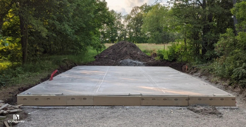 Why Your Steel Building Cost Is Higher Than Expected? (Full Breakdown With Hidden Fees) 2 Freshly poured concrete slab foundation in a clearing surrounded by trees, prepared for a future steel building installation.