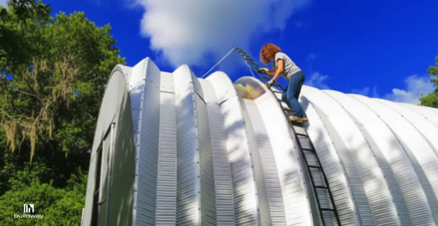Thinking About Quonset Buildings? Facts, Myths, and Real-World Uses 3 A woman on a ladder cleaning the curved metal roof of a building under a bright blue sky, surrounded by lush green trees.